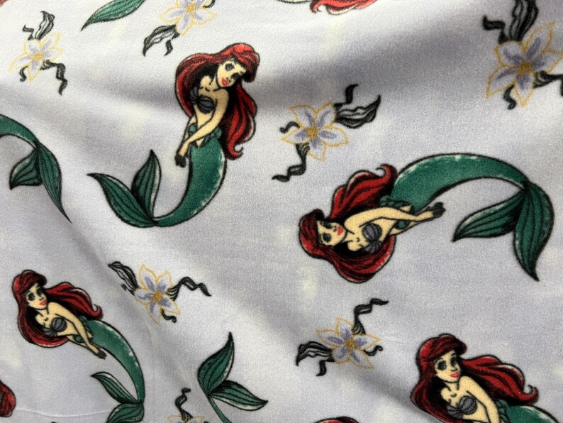 May include: A white fabric with a repeating pattern of a mermaid with red hair and a green tail. The mermaid is holding a seashell in her hand. There are also white flowers with yellow centers in the pattern.