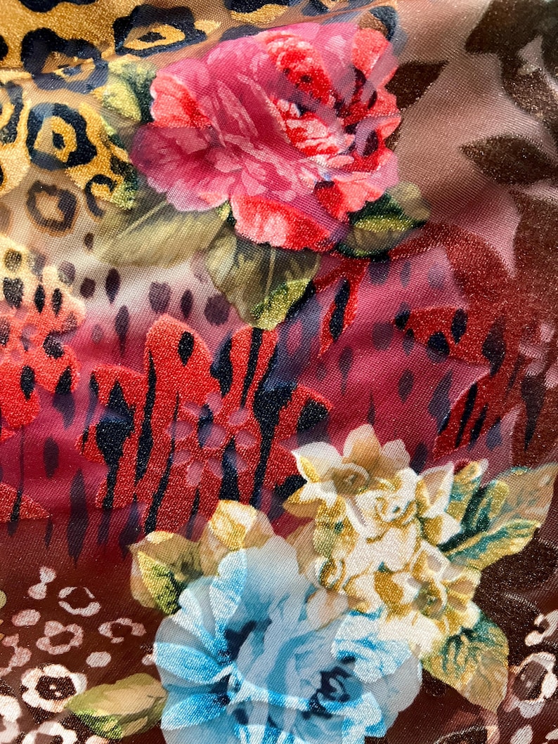 May include: A close-up of a fabric with a floral and animal print. The fabric features a large pink rose, a smaller blue rose, and a leopard print pattern. The fabric is a mix of red, blue, pink, yellow, and brown.