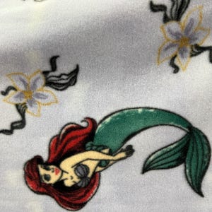 May include: A white fabric with a repeating pattern of a mermaid with red hair and a green tail, and a white flower with yellow accents.