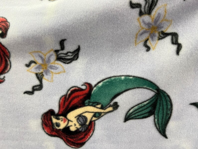 May include: A white fabric with a repeating pattern of a mermaid with red hair and a green tail, and a white flower with yellow accents.