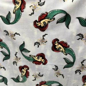 May include: A white fabric with a repeating pattern of Ariel, the Little Mermaid, as a mermaid. She is depicted with red hair, a green tail, and a seashell bra. The background is a light blue with white flowers.