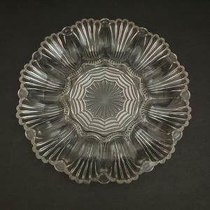 Peut inclure: Plat de service en verre transparent avec un bord festonné et un motif en étoile au centre. Le plat présente un motif radial de rainures s'étendant du centre au bord. Le plat est rond et repose sur une surface sombre.