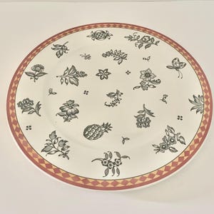 Puede incluir: Un plato redondo de color crema con un borde decorativo y motivos florales y de frutas. El borde es una mezcla de diamantes rojos y amarillos. El plato presenta diseños florales y de piña en negro y gris.