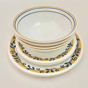 Puede incluir: Un conjunto de cuencos y platos de cerámica blanca. Los cuencos tienen un borde azul y dorado. Los platos tienen un borde dorado con un diseño de hojas azules. El conjunto está sobre una superficie blanca.