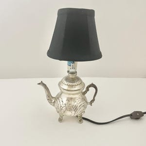 Może przedstawiać: Lampa stołowa z podstawą w kształcie srebrnego metalowego czajnika, ozdobioną tłoczonymi wzorami kwiatowymi, trzema małymi nóżkami i zakrzywionym uchwytem. Lampa posiada jednolity czarny abażur z tkaniny oraz czarny przewód zasilający z przełącznikiem.