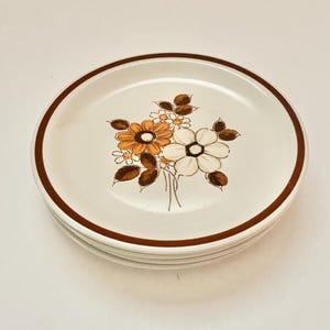 May include: A stack of four white ceramic plates with a brown rim. Each plate features a floral design in shades of orange, white, and brown. The plates are stacked on top of each other.