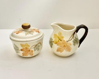 May include: A cream-coloured ceramic sugar bowl with a matching lid and a creamer pitcher. Both pieces feature a painted pattern of orange, yellow, and green autumn leaves. The creamer has a dark brown handle, and the lid has a brown knob.