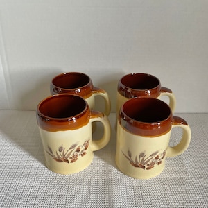 May include: Four ceramic beer steins. Each stein has a cream-coloured body with a brown rim and handle. A wheat design is printed on the front. The interior of the steins is brown.