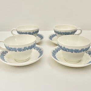 May include: Four white teacups and saucers with a light blue floral design. The cups have handles and the saucers are round. The blue design is around the rim of the cups and saucers. The teacups and saucers are on a white surface.