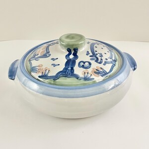 以下が含まれることがあります： 蓋付きのセラミック製サービングディッシュ。ディッシュは白で、青い縁とハンドルが付いています。蓋は白で、青、緑、桃色でペイントされたデザインが施されています。蓋には緑色のノブが付いています。
