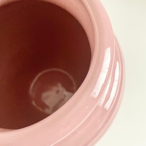 May include: A pink ceramic jar with a smooth, glossy finish. The jar has a wide opening and a rounded shape.