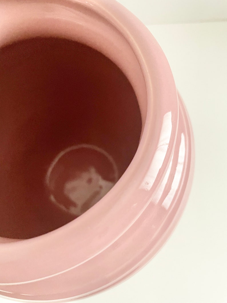 May include: A pink ceramic jar with a smooth, glossy finish. The jar has a wide opening and a rounded shape.