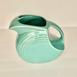 May include: A turquoise ceramic pitcher with a rounded body and a wide, flared spout. The pitcher has a smooth, glossy finish and a simple, elegant design.