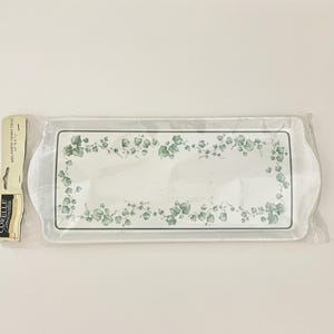 May include: A rectangular white melamine tray featuring a green ivy leaf border pattern. The tray is sealed in clear plastic packaging with a Corelle Coordinates tag that lists the dimensions as 15 inches by 6 inches.