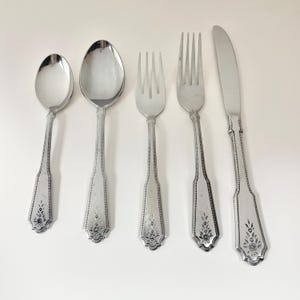 May include: A five-piece silver-toned flatware set consisting of a small spoon, a large spoon, a small fork, a large fork, and a dinner knife. Each piece features a decorative floral engraving at the base of the handle and a beaded border design.