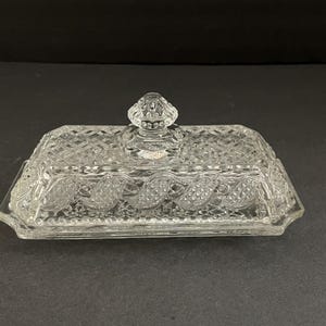 Pode incluir: Uma manteigueira retangular de vidro transparente com um padrão de diamante e uma tampa decorativa. A tampa tem um botão para facilitar a elevação. O prato tem uma borda ondulada e um design detalhado.