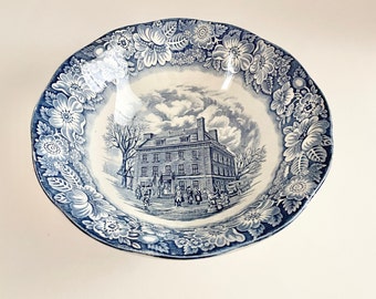 May include: A blue and white ceramic bowl with a floral border and a central scene of a building with people. The bowl has a scalloped edge and a glossy finish. The floral border features large, stylized flowers.