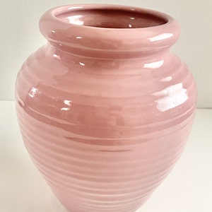 May include: A pale pink ceramic vase with a ribbed design. The vase has a rounded bottom and a wide opening.