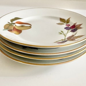 May include: A stack of four white ceramic plates with a gold rim. Each plate features a different fruit design, including pears and blackberries. The plates are stacked on top of each other.