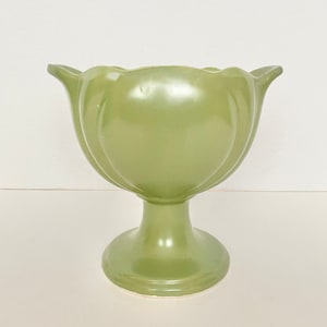 May include: A green ceramic planter with a wide, scalloped rim and a pedestal base. The planter has a smooth, glossy finish.