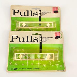 Op de afbeelding: Twee vintage kastgrepen op hun originele verpakking. De grepen zijn goudkleurig met ruitvormige accenten. De verpakking is groen en wit met het woord "Pulls" in grote zwarte letters. De tekst "California Styled Hardware" is ook aanwezig.