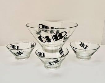 Puede incluir: Un juego de cuatro cuencos de vidrio transparente para patatas fritas y salsas. El cuenco más grande tiene la palabra "CHIP" en letras negras. Los cuencos más pequeños tienen la palabra "DIP" en letras negras. Los cuencos están dispuestos en forma de nido.