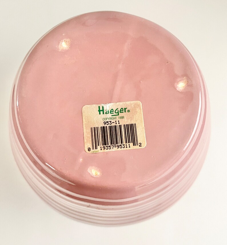 May include: The bottom of a pink ceramic vase with white stripes. The vase has a green label with the text "Haeger" and "Copyright 1998" and the number "953-11". A barcode is also on the label.