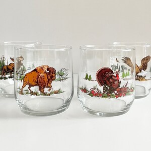 May include: Set of four clear glass tumblers with a winter scene design. Each tumbler features a different animal: a moose, a buffalo, a turkey, and an eagle. The animals are depicted in a snowy landscape with trees and other foliage.