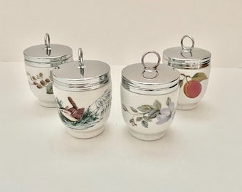 May include: Four white porcelain egg cups with silver-colored lids. Each cup features a different hand-painted design, including birds, flowers, and fruit. The lids have a small ring handle. A charming set for serving eggs.