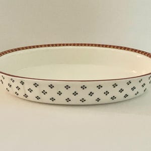 Op de afbeelding: Ovale keramische serveerschaal met een wit interieur en een decoratieve rand. De rand heeft een bruin geometrisch patroon en de buitenkant heeft een herhalend patroon van kleine, donkerblauwe bloemmotieven. Een klassiek stuk om te serveren.