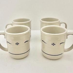 May include: A set of four off-white ceramic mugs featuring a dark blue decorative band with a central floral motif and a single horizontal line near the base. Each mug has a sturdy handle and a smooth, rounded rim, arranged in a two-by-two grid.