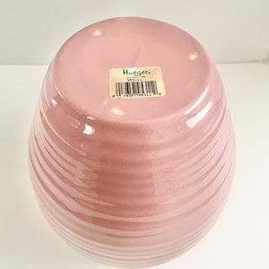 May include: A pink ceramic vase with a ribbed design. The bottom of the vase has a green sticker with the text "Hueger" and a barcode.