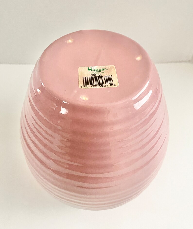 May include: A pink ceramic vase with a ribbed design. The bottom of the vase has a green sticker with the text "Hueger" and a barcode.