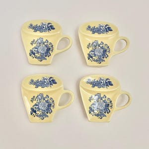 May include: Four yellow ceramic cup-shaped coasters with blue floral designs. Each coaster has a handle and a flat top, resembling a teacup. The floral pattern includes roses and other flowers.