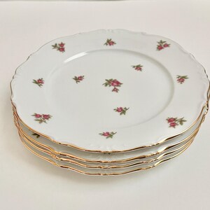 May include: A set of four white porcelain dinner plates with a delicate floral pattern and gold trim. The plates have a scalloped edge and are stacked on top of each other.