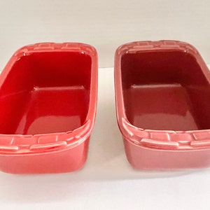 以下が含まれることがあります： テクスチャ加工された隆起した縁のある赤いセラミック製のパン型が2つ。左側の型は右側の型よりも明るい赤色です。