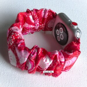 May include: A red and white patterned fabric watch band with glitter. The band is wrapped around a silver Apple Watch with a black face and a red button.