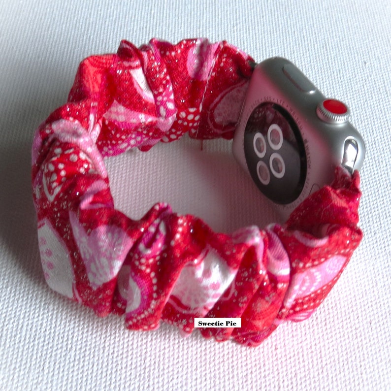 May include: A red and white patterned fabric watch band with glitter. The band is wrapped around a silver Apple Watch with a black face and a red button.