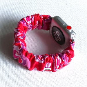 May include: A pink and purple patterned fabric Apple Watch band with a silver Apple Watch. The band is made of a stretchy fabric and has a ruffled look. The watch face is black and has a red button on the side. The text "Amore" is visible on the band.