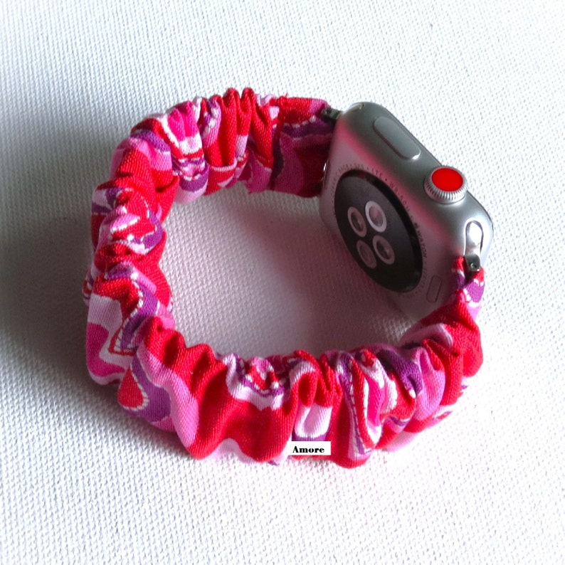 May include: A pink and purple patterned fabric Apple Watch band with a silver Apple Watch. The band is made of a stretchy fabric and has a ruffled look. The watch face is black and has a red button on the side. The text "Amore" is visible on the band.