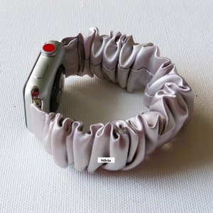May include: A silver Apple Watch with a light gray, silky, elastic band. The band is made of gathered fabric and has the word "Silvia" printed on it.