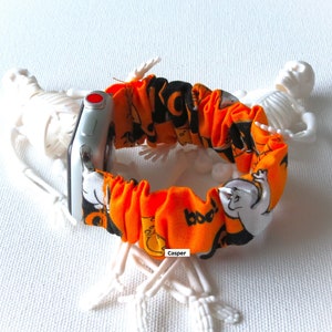 Peut inclure: Bracelet de montre orange et noir sur le thème d'Halloween avec un fantôme blanc et le mot "boo!" imprimé dessus. Le bracelet est en tissu et est réglable. Le bracelet de montre est présenté sur une surface blanche avec deux squelettes en plastique.