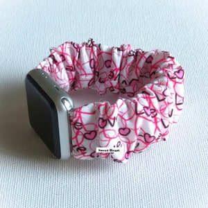 May include: White Apple Watch band with a pink and white heart pattern. The band is made of stretchy fabric and has the words "Sweet Heart" printed on it.