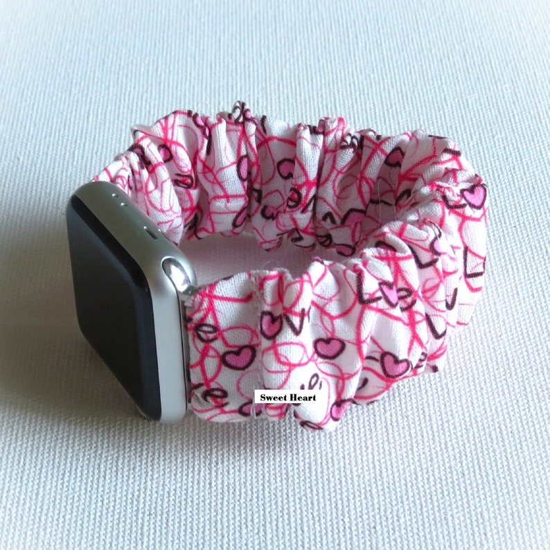 May include: White Apple Watch band with a pink and white heart pattern. The band is made of stretchy fabric and has the words "Sweet Heart" printed on it.