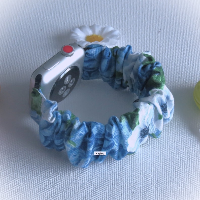 May include: A silver Apple Watch with a blue and white floral fabric band. The band is a scrunchie style and has a white daisy flower on top.