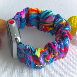 May include: A colorful scrunchie watch band with a vibrant, abstract pattern in shades of blue, pink, red, and yellow. The band is attached to a silver smartwatch with a red button. The word "Astralea" is printed on the band.