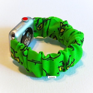 May include: A silver Apple Watch with a red button is attached to a green fabric watch band. The band is patterned with a Grinch face design in black and yellow. The word "Grinch" is printed on the band.