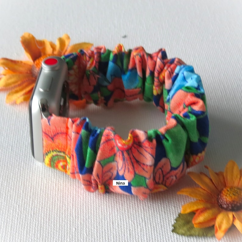 May include: A colorful floral patterned fabric watch band for an Apple Watch. The band is made of a stretchy fabric and has a bright orange, green, blue, and pink color scheme. The band is shown on a silver Apple Watch.