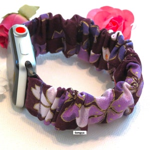 K&ouml;nnte beinhalten: Lila und wei&szlig;es Blumen-Apple Watch-Armband mit goldener Akzent. Das Armband besteht aus einem weichen, dehnbaren Stoff und ist so konzipiert, dass es eng am Handgelenk sitzt.