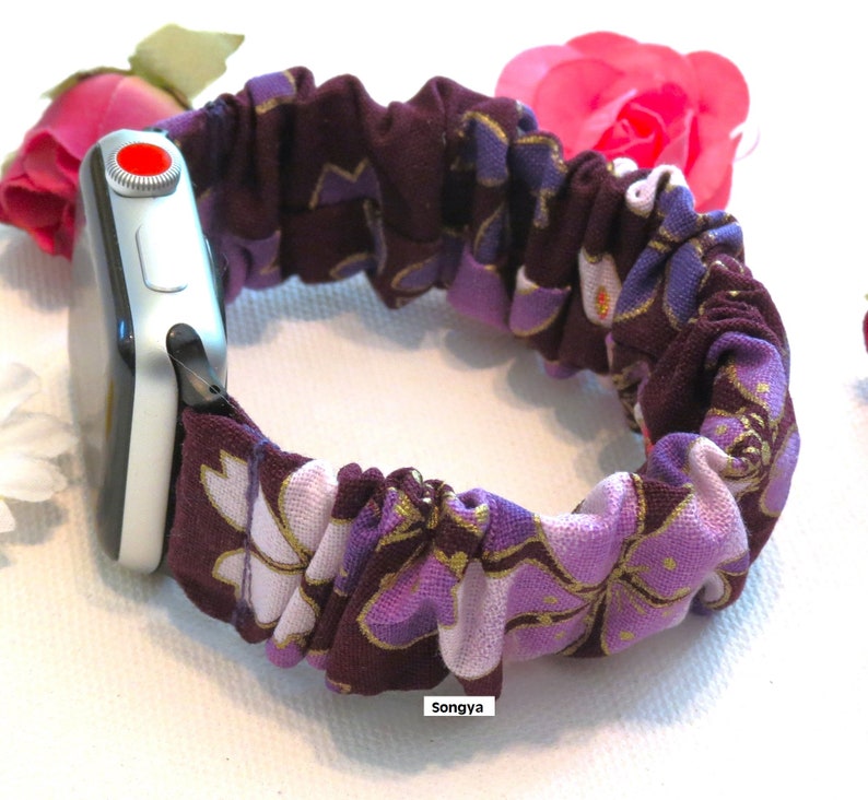 K&ouml;nnte beinhalten: Lila und wei&szlig;es Blumen-Apple Watch-Armband mit goldener Akzent. Das Armband besteht aus einem weichen, dehnbaren Stoff und ist so konzipiert, dass es eng am Handgelenk sitzt.